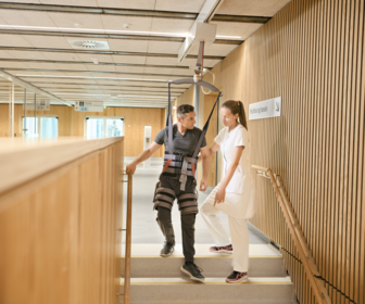 Patiënt in Gait Trainer tilband ondersteund door GH3+ plafondlift en Trainer Module – ontvangt instructies van zorgverlener tijdens veilige trapafgang bij neurorevalidatie in Rigshospitalet.