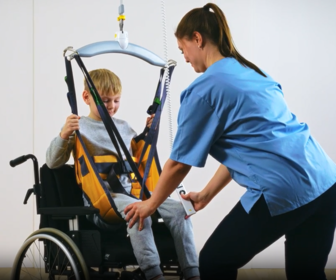 Child being transferred from a wheelchair using an Active Micro Plus Kids sling and ceiling hoist in an instruction video.